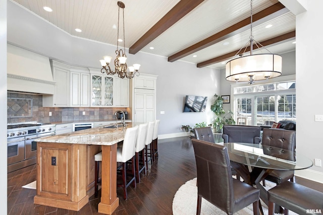 kitchen with a kitchen island with sink, premium range hood, range with two ovens, light stone counters, and white cabinetry