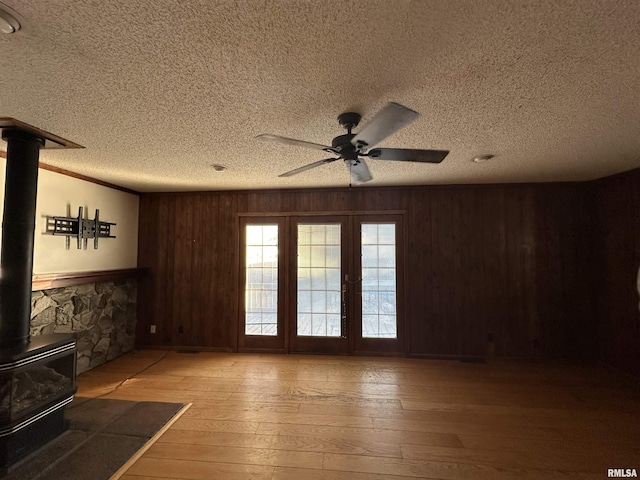 unfurnished living room with ceiling fan, wood-type flooring, wooden walls, a wood stove, and a textured ceiling