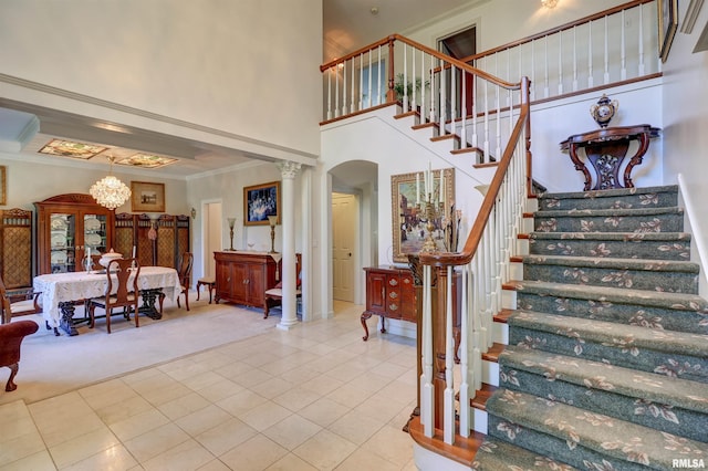 stairs featuring ornate columns, a towering ceiling, tile patterned flooring, crown molding, and an inviting chandelier