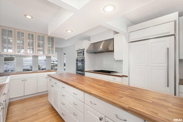 kitchen featuring wall chimney exhaust hood, butcher block counters, white cabinetry, appliances with stainless steel finishes, and decorative backsplash