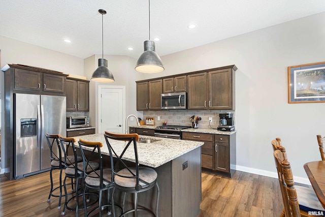 kitchen featuring appliances with stainless steel finishes, a kitchen island with sink, light stone counters, tasteful backsplash, and decorative light fixtures