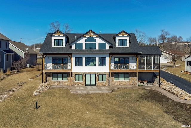 back of house featuring stone siding, a shingled roof, a lawn, and a sunroom