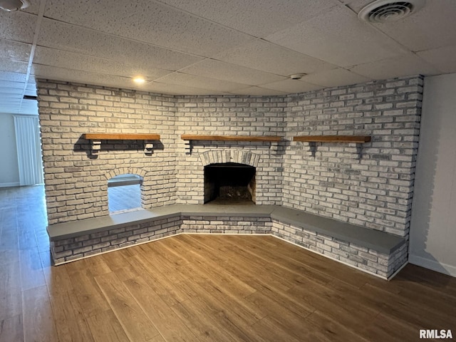 unfurnished living room with a paneled ceiling, wood-type flooring, brick wall, and a brick fireplace