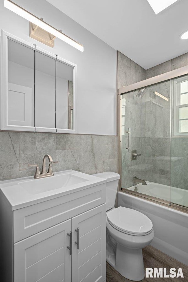 full bathroom featuring toilet, wood-type flooring, bath / shower combo with glass door, tile walls, and vanity