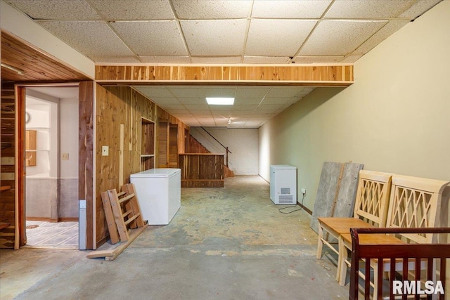 basement featuring a paneled ceiling, wooden walls, and refrigerator