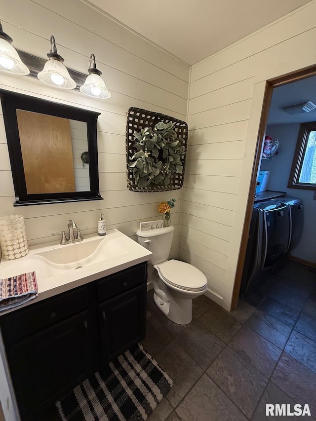 half bath featuring wooden walls, washer and clothes dryer, toilet, stone tile flooring, and vanity