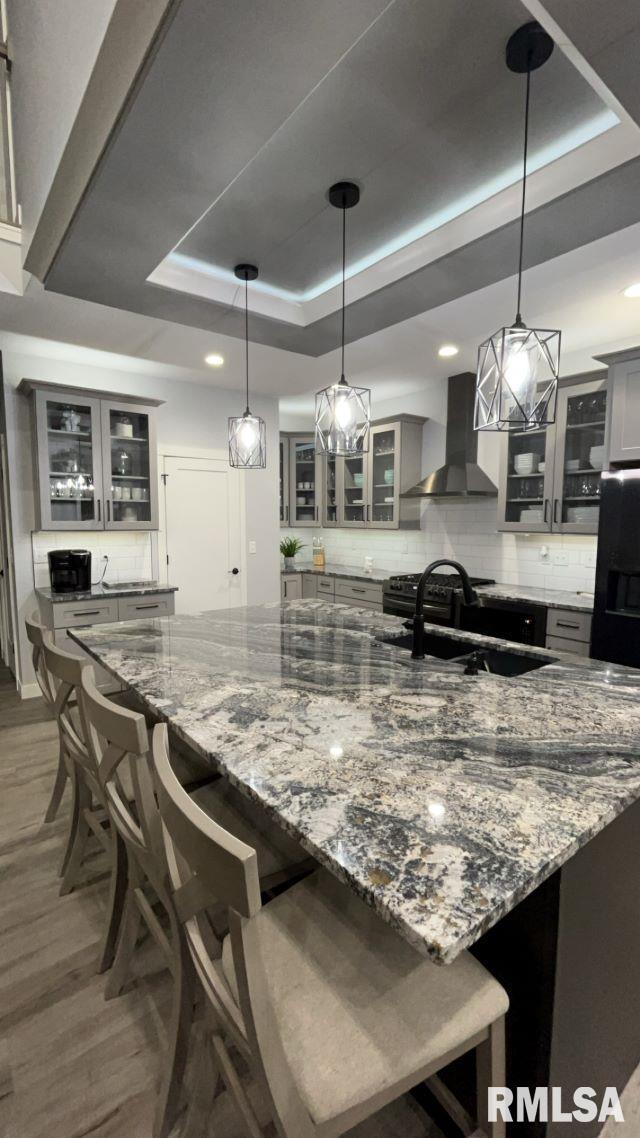 kitchen featuring light stone countertops, a tray ceiling, a breakfast bar, and wall chimney exhaust hood