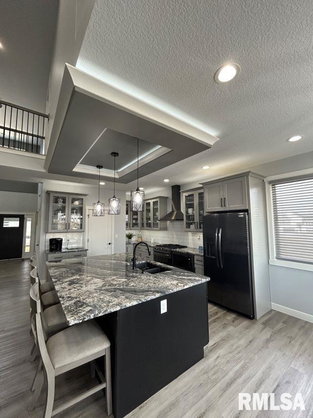 kitchen featuring hanging light fixtures, a raised ceiling, black fridge, light wood-type flooring, and wall chimney exhaust hood