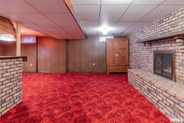 basement featuring carpet floors, a brick fireplace, wood walls, and a drop ceiling