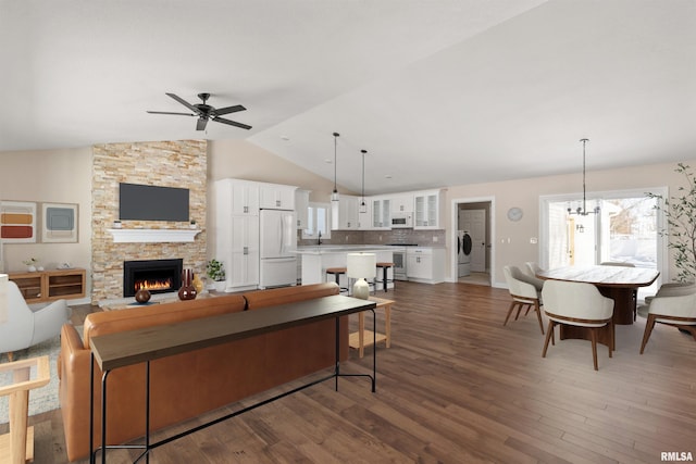 living area with lofted ceiling, a stone fireplace, dark wood-type flooring, and ceiling fan with notable chandelier