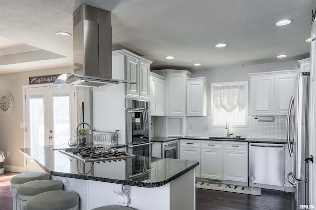 kitchen featuring island range hood, dark stone counters, stainless steel appliances, white cabinetry, and a sink