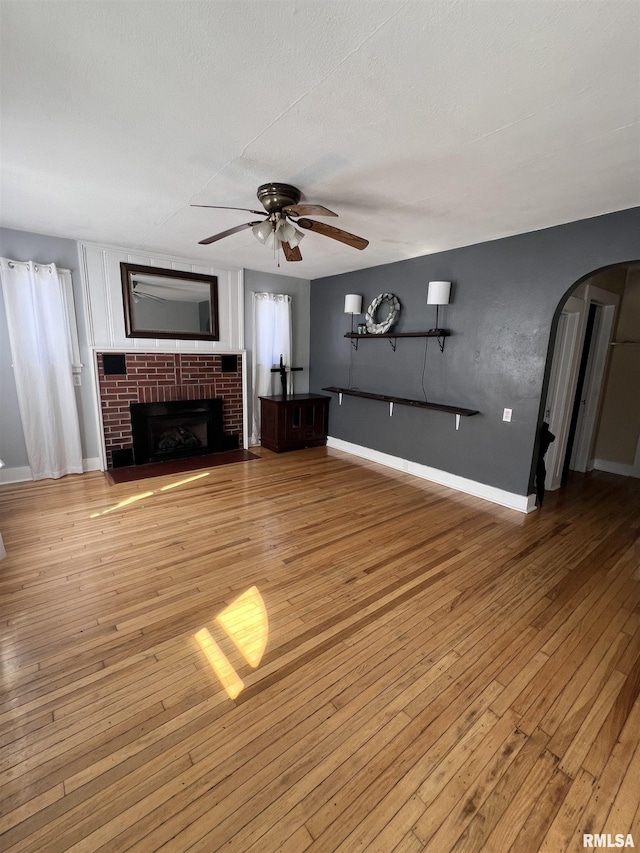 unfurnished living room featuring a ceiling fan, arched walkways, a brick fireplace, and light wood-style flooring