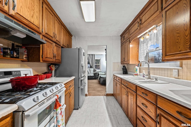 kitchen featuring under cabinet range hood, a sink, backsplash, brown cabinets, and gas range oven