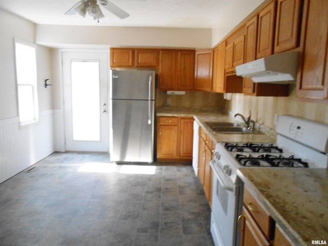 kitchen featuring under cabinet range hood, gas range gas stove, a sink, freestanding refrigerator, and brown cabinetry