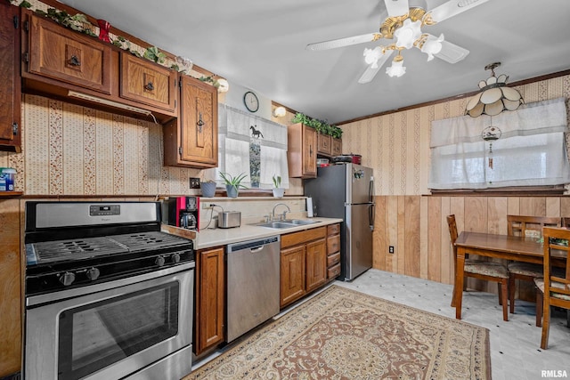 kitchen featuring a wainscoted wall, brown cabinets, a sink, stainless steel appliances, and wallpapered walls