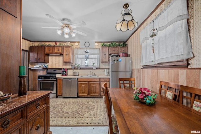 kitchen with wallpapered walls, wainscoting, appliances with stainless steel finishes, brown cabinets, and a sink