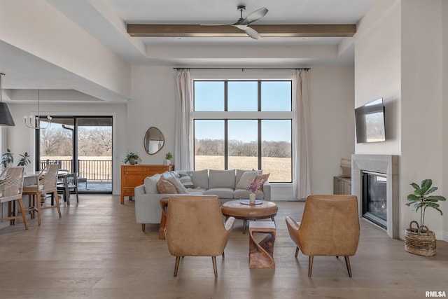 living area with light wood-type flooring, beam ceiling, a towering ceiling, a glass covered fireplace, and a ceiling fan