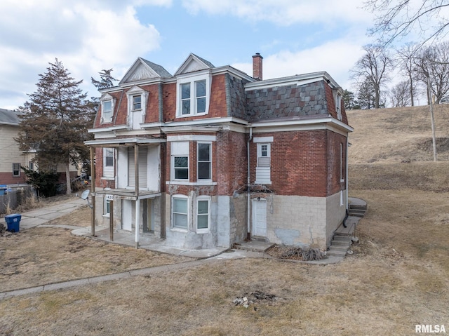 view of front of house featuring mansard roof, a chimney, a shingled roof, and brick siding