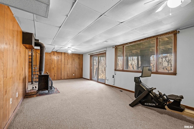 workout area featuring wooden walls, ceiling fan, a wood stove, carpet, and a paneled ceiling