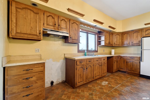 kitchen featuring under cabinet range hood, light countertops, brown cabinets, freestanding refrigerator, and a sink