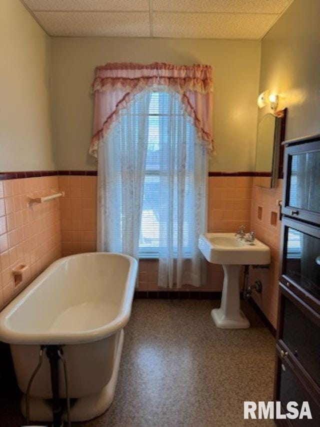 bathroom with a wainscoted wall, a paneled ceiling, a soaking tub, and tile walls