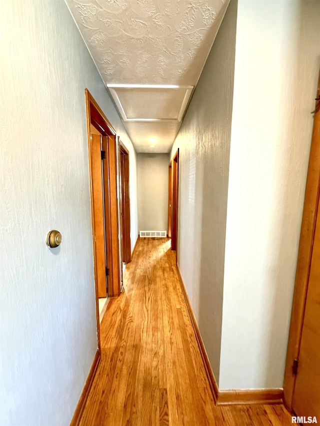 hall featuring a textured ceiling, visible vents, baseboards, light wood finished floors, and attic access