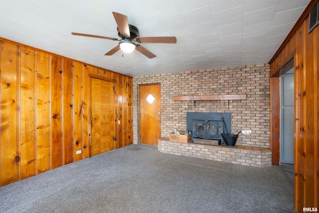 unfurnished living room with a wood stove, wood walls, a ceiling fan, and carpet flooring