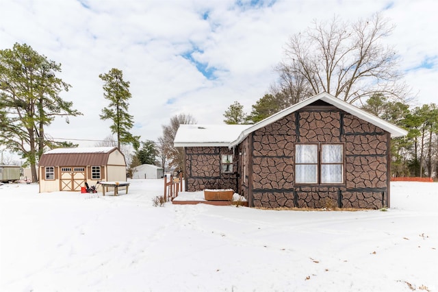 snow covered rear of property with an outbuilding, stone siding, a shed, and metal roof