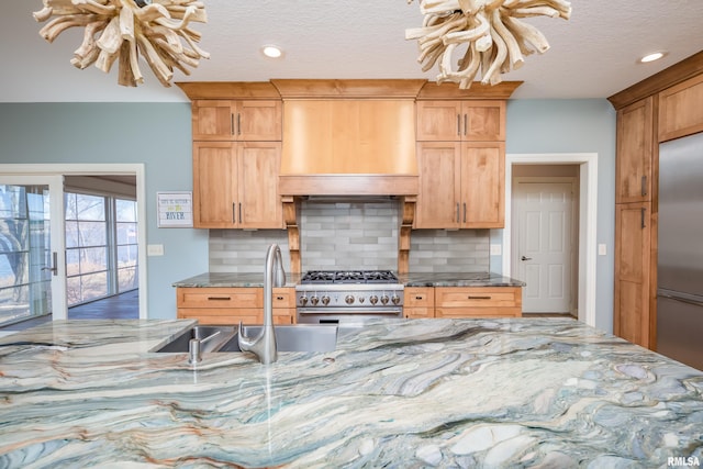 kitchen featuring premium appliances, a sink, backsplash, wall chimney exhaust hood, and dark stone countertops