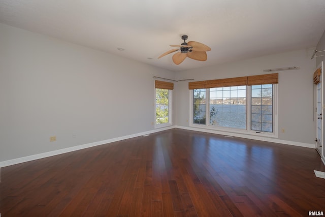 empty room with a water view, dark wood finished floors, a ceiling fan, and baseboards