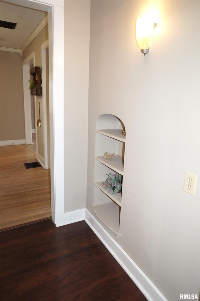 hallway with built in shelves, dark wood-style flooring, crown molding, visible vents, and baseboards