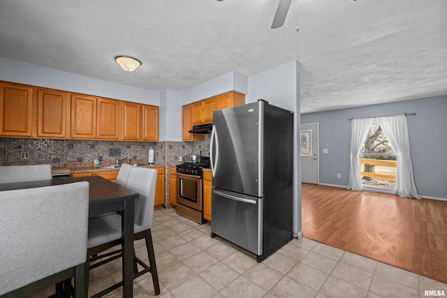 kitchen featuring baseboards, appliances with stainless steel finishes, brown cabinets, under cabinet range hood, and backsplash