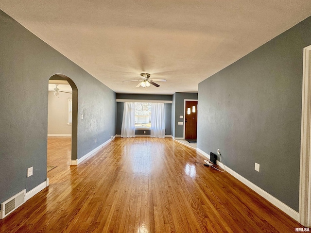 unfurnished living room featuring a ceiling fan, wood finished floors, visible vents, baseboards, and arched walkways