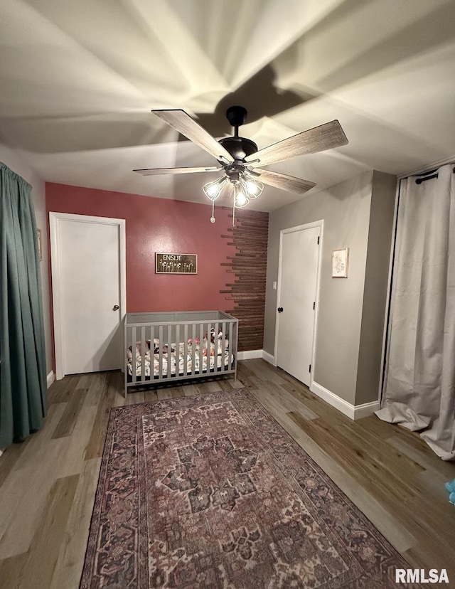 unfurnished bedroom featuring a crib, wood finished floors, baseboards, and ceiling fan