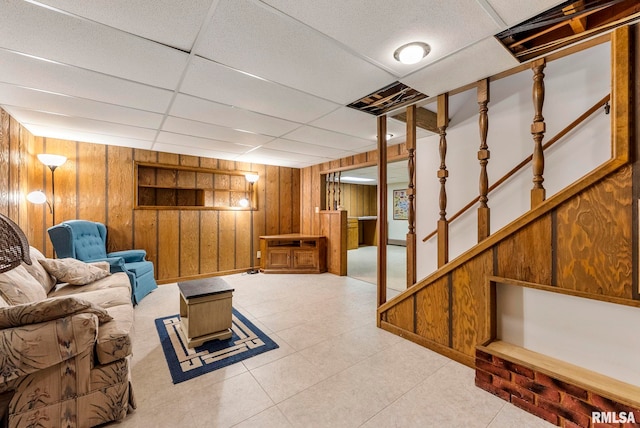 tiled living room featuring a drop ceiling, wood walls, and stairs