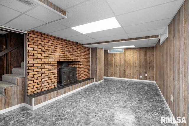 finished basement featuring stairway, wooden walls, visible vents, a wood stove, and a paneled ceiling