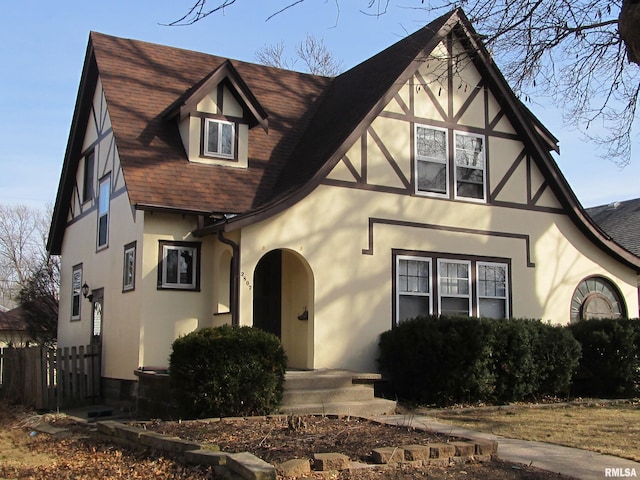 english style home featuring fence, roof with shingles, and stucco siding