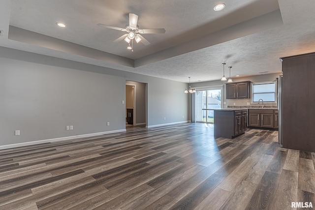 unfurnished living room featuring dark wood-type flooring, ceiling fan with notable chandelier, a sink, a tray ceiling, and baseboards