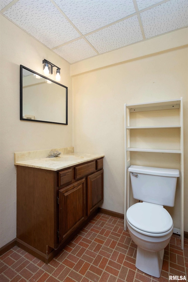 bathroom featuring brick floor, a drop ceiling, baseboards, and vanity