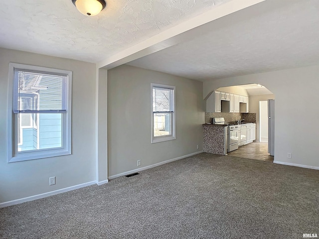 unfurnished living room with visible vents, a textured ceiling, arched walkways, dark colored carpet, and baseboards