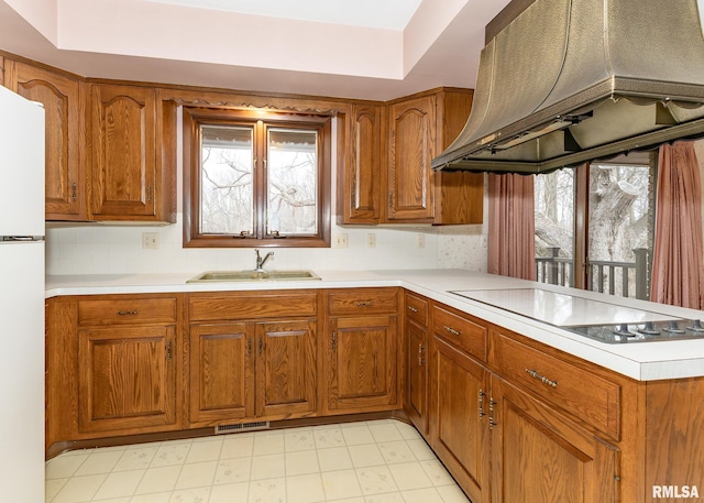 kitchen with electric stovetop, freestanding refrigerator, exhaust hood, brown cabinetry, and a sink