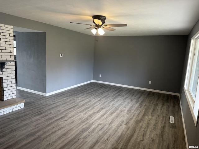 spare room featuring dark wood-style flooring, visible vents, a brick fireplace, baseboards, and ceiling fan