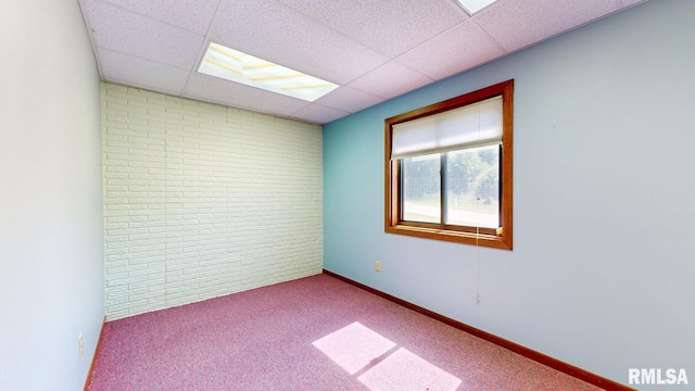 carpeted empty room featuring brick wall, baseboards, and a drop ceiling