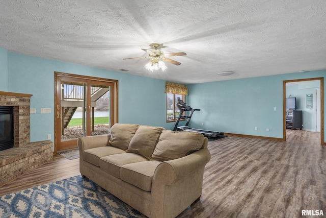 living area with wood finished floors, ceiling fan, baseboards, a textured ceiling, and a brick fireplace
