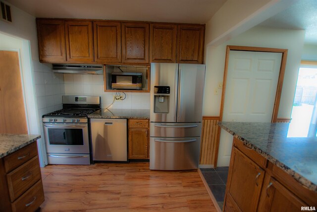 kitchen with stone countertops, tasteful backsplash, under cabinet range hood, brown cabinets, and stainless steel appliances