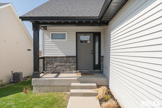 view of exterior entry with a yard, roof with shingles, stone siding, and cooling unit