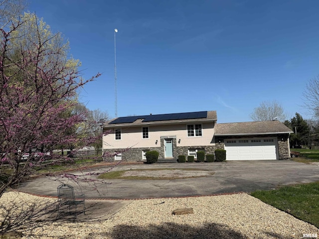 raised ranch featuring roof mounted solar panels, driveway, stone siding, and an attached garage