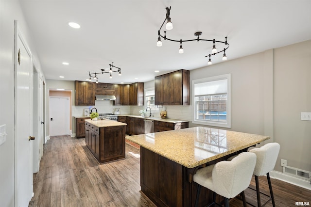 kitchen featuring stainless steel dishwasher, a center island, dark brown cabinetry, under cabinet range hood, and rail lighting