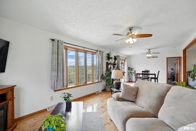 living room with light wood-style floors, a glass covered fireplace, a ceiling fan, visible vents, and baseboards