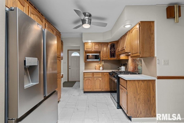 kitchen featuring ceiling fan, stainless steel appliances, light countertops, marble finish floor, and brown cabinetry